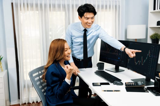 Successful smiling Asian business partners raising fist up together in profitable project job in stock exchange database on pc wearing formal suit at modern home office at side view image. Infobahn.