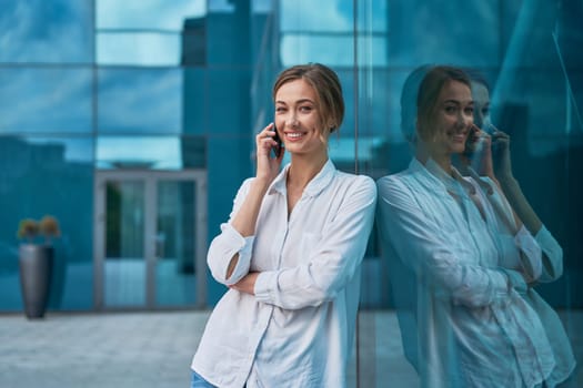 Portrait of smiling young businesswoman in white shirt talking on smartphone while leaning on glass office building. Confident female entrepreneur discussing on cellphone outside.