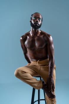 Portrait of serious gay man with eyeshadow sitting on stool. Confident muscular bearded individual against blue background. African American gay person with bright makeup.