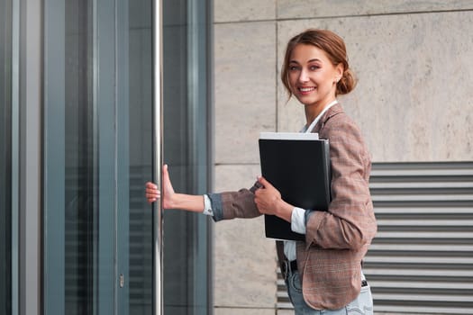 Portrait of smiling young businesswoman holding folder and entering corporate office. Confident female entrepreneur opening the door, symbolizing the concept of new opportunities.