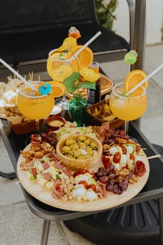 A variety of aperitif snacks on wooden round dishes with colorful alcoholic cocktails on a garden table under an umbrella in the backyard of a house on a spring sunny day, side view close-up.