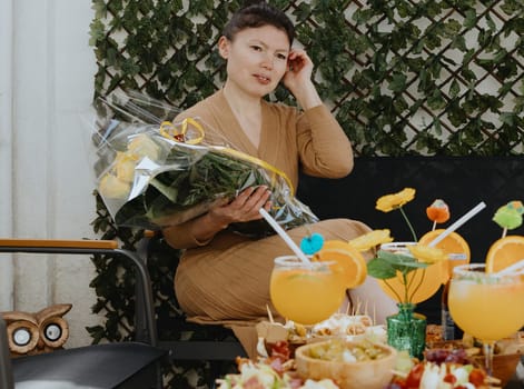 Portrait of one young beautiful Caucasian brunette girl in a knitted dress holding a bouquet of yellow roses in her hands, sitting on a garden chair in the backyard of a house on a spring sunny day and thoughtfully looking to the side, straightening her hair with her hand, celebrating her birthday, side view close-up.