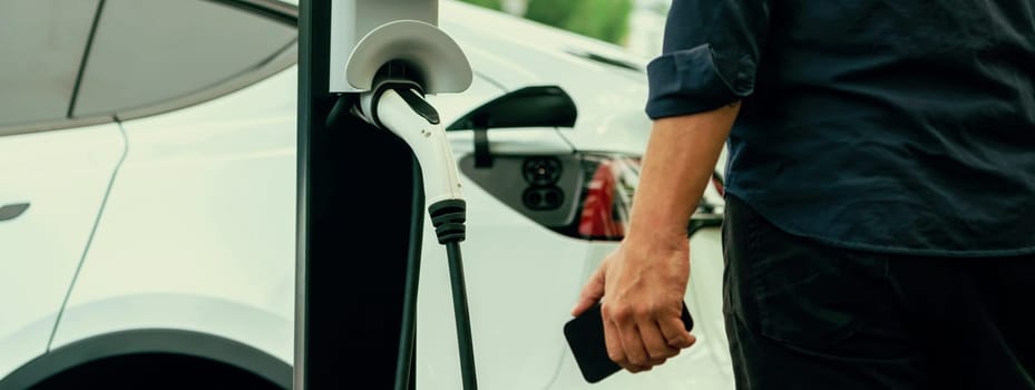 Man using smartphone online banking application to pay for electric car battery charging from EV charging station during vacation road trip at national park or summer forest. Panorama Exalt