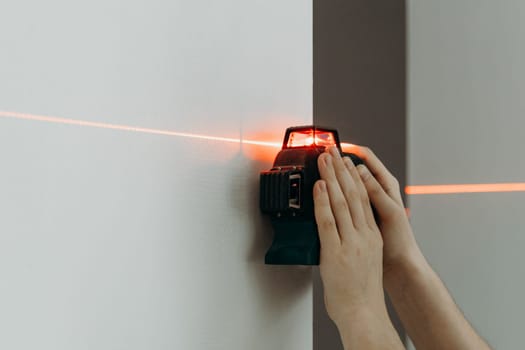 One young unrecognizable Caucasian guy holds with both hands, using a modern laser level to measure a straight line on the wall, before drilling with a drill, side view close-up with small copy space on the left.