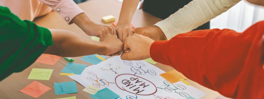 Young happy creative startup group join circle fist bump hands together surrounded by marketing strategy mind map and colorful sticky notes at meeting room. Unity and teamwork concept. Variegated.