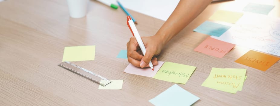 Closeup imaged of young creative businesswoman writes on colorful sticky note and places it on brainstorming mind map at meeting room. Creative and teamwork concept. Focus on hand. Variegated.