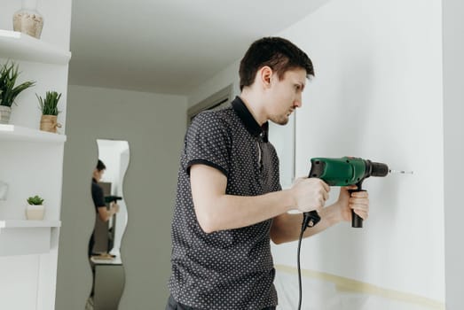 One young handsome Caucasian brunette guy stands sideways, holds a drill with both hands and drills a hole in a white wall, standing in a room and reflected behind in a figured mirror on a spring day, side view close-up with depth of field.