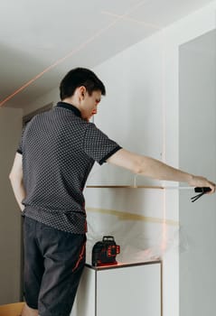 One young handsome Caucasian brunette guy stands half sideways, holds a measuring construction tape in his hands and checks the evenness of a hung wooden shelf on a white wall in a room on a spring day using a laser level, side view close-up with depth of field.