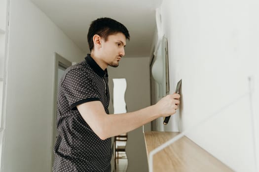 One young handsome Caucasian brunette guy stands half sideways and covers a screw hole on a white wall with a special putty using a spatula, standing in a room on a spring day, close-up side view with depth of field.