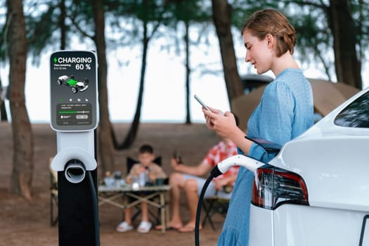 Outdoor adventure and family vacation camping at sea travel by eco friendly car. Woman or mother check car's battery with smartphone while charging EV car frin charging station in campsite. Perpetual