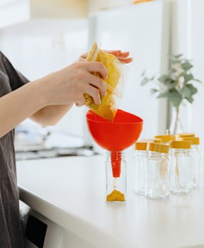 One young Caucasian unrecognizable girl pours yellow seasoning spice from a transparent bag into a glass jar while standing at a white table in the kitchen on a summer day, side view close-up with depth of field.