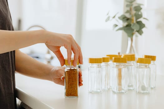 One young Caucasian unrecognizable girl closes with her hands a wooden lid mix seasoning spice for fish in a glass jar, standing at a white table in the kitchen on a summer day, side view close-up with depth of field.