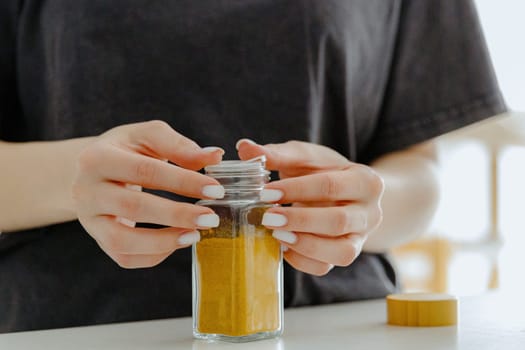 One young Caucasian unrecognizable girl closes the lid of a glass jar with curry spice while standing at a white table in the kitchen on a summer day, side view close-up with depth of field. Eco-friendly storage concept.