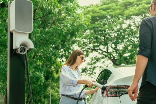 Lovely young couple wearing sun glasses recharging battery for electric car during road trip travel EV car in natural forest or national park. Eco friendly travel during vacation and holiday. Exalt