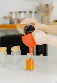 One young Caucasian unrecognizable girl pours seasoning spice smoked ground paprika from a plastic jar into a glass transparent jar, standing at a white table in the kitchen on a summer day, using a red watering can, bottom side view close-up with depth of field. Eco-friendly storage concept.