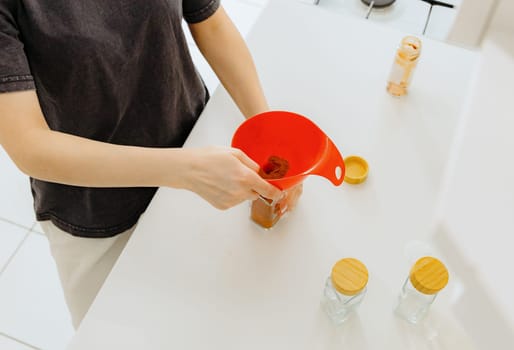 One young Caucasian unrecognizable girl pours seasoning spice ground paprika into a new glass transparent jar, standing at a white table in the kitchen on a summer day, using a red watering can, top view close-up with depth of field. Eco-friendly storage concept.