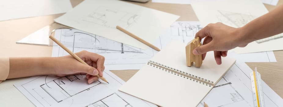 Professional architect hand draws a blueprint on table with architectural document and wooden block scatter around at office. Design and Planing concept. Focus on hand. Closeup. Delineation.