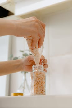 One young Caucasian unrecognizable girl pours pink salt seasoning spice from a plastic bag into a new glass transparent jar, standing at a white table in the kitchen on a summer day, bottom side view close-up with depth of field. Eco-friendly storage concept.