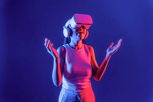 Smart female stand with surrounded by cyberpunk neon light wear VR headset connecting metaverse, futuristic cyberspace community technology. Elegant woman look generated virtual area. Hallucination.