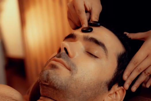 Caucasian man enjoying relaxing anti-stress head massage with hot stone and pampering facial beauty skin recreation leisure in warm candle light ambient salon spa in resort or hotel. Quiescent