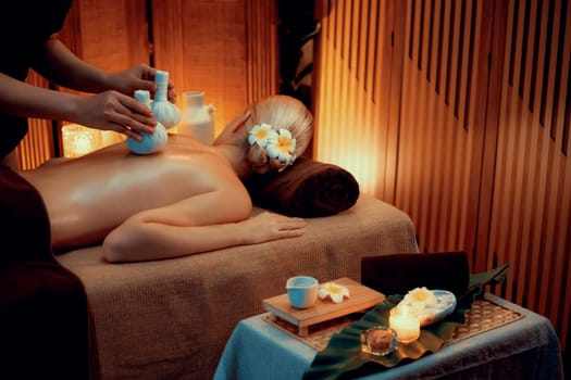 Hot herbal ball spa massage body treatment, masseur gently compresses herb bag on woman body. Tranquil and serenity of aromatherapy recreation in warm lighting of candles at spa salon. Quiescent