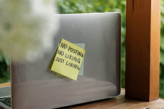 Paper note with text NO POSTING NO LIKING JUST LIVING in laptop garden background. Concept of digital detox, unplugging from technology break. Mindful living without gadgets