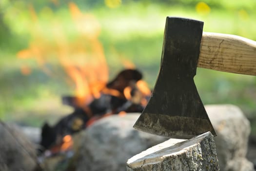 Closeup view of axe with a wooden handle embedded in a log of wood, with a campfire in the background