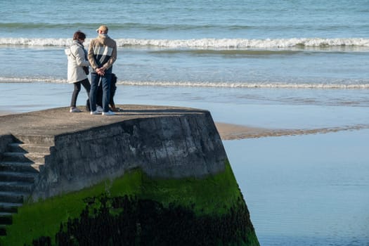 Unrecognizable couple and dog on Normandy France Gold beach peir, seawall and promenade. View of sand, blue sea and rock seawall. High quality photo