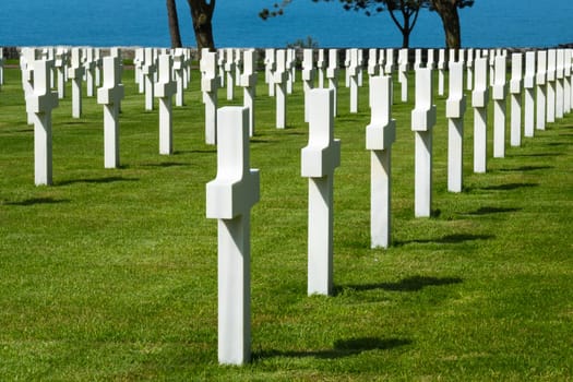 American cemetery at Normandy area. Grave with blue ocean in foreground at WWII memorial. Lines of grave stones on green grass. High quality photo