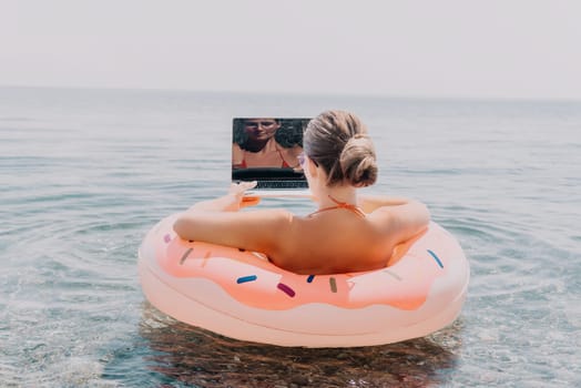 Woman freelancer works on laptop swimming in sea on pink inflatable ring. Pretty lady typing on computer while floating in the sea on inflatable donut at sunset. Freelance, remote work on vacation