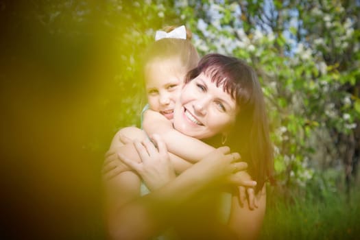 Happy mother and daughter enjoying rest, playing and fun on nature on a green lawn with dandelions and blooming apple tree on background. Woman and girl resting outdoors in summer and spring day