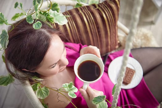Middle aged woman relaxing and Enjoying Coffee in Cozy Indoor Swing in living room. Mature female person resting alone. A break in work