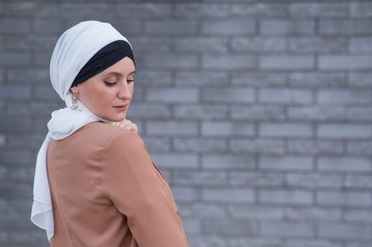 Portrait of a young blue-eyed woman in a hijab against a gray brick wall. A Muslim woman looks at the camera turning around