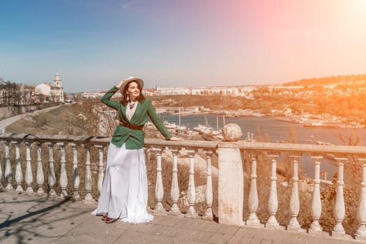 Woman walks around the city, lifestyle. Happy woman in a green jacket, white skirt and hat is sitting on a white fence with balusters overlooking the sea bay and the city