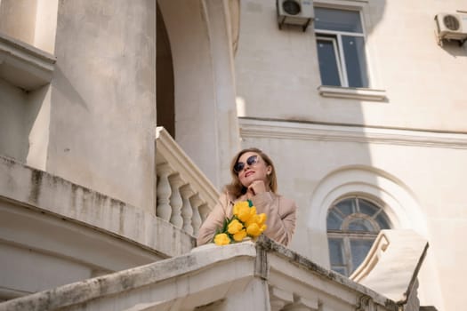 A woman wearing sunglasses and holding a bouquet of yellow flowers stands on a balcony. The scene is peaceful and serene, with the woman looking out over the city