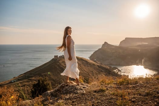 A woman in a white dress stands on a hill overlooking the ocean. The scene is serene and peaceful, with the woman's dress billowing in the wind. The combination of the ocean