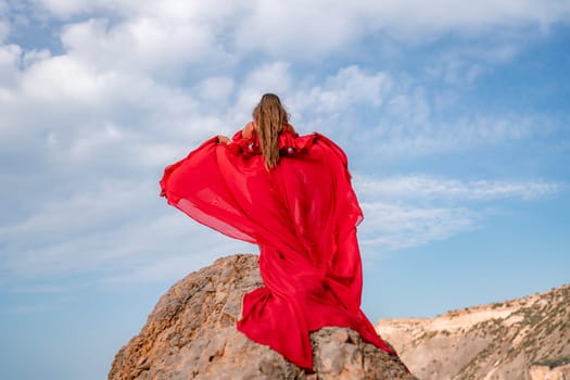 woman sky red dress. Woman with long hair on a sunny seashore in a red flowing dress, back view, silk fabric waving in the wind. Against the backdrop of the blue sky and mountains on the seashore