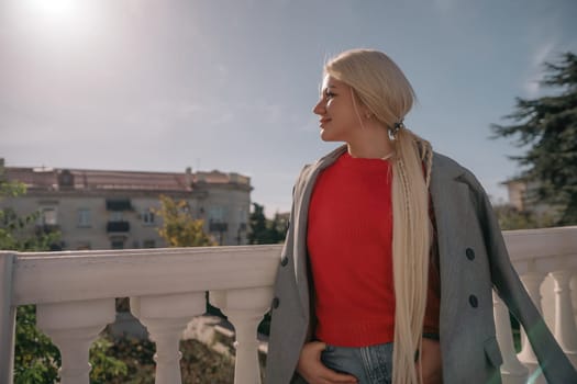 A blonde woman wearing a red shirt and gray jacket stands on a balcony. She is wearing ripped jeans and has her hands in her pockets