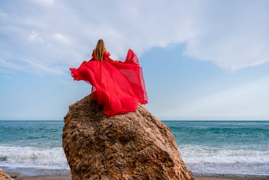 woman sea red dress. Woman with long hair on a sunny seashore in a red flowing dress, back view, silk fabric waving in the wind. Against the backdrop of the blue sky and mountains on the seashore