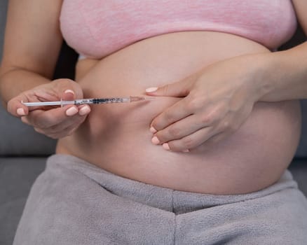 A pregnant woman puts an injection of insulin while sitting on the couch. Close up of the belly