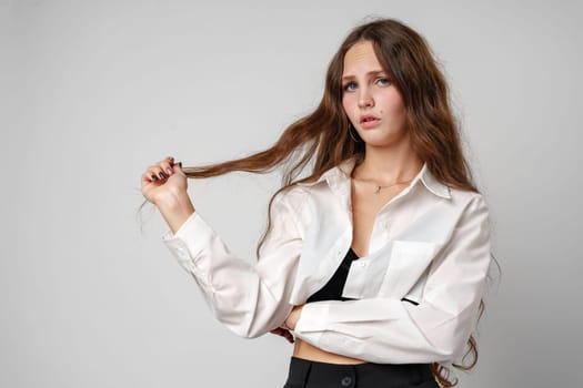 A young woman stands against a plain background, her gaze fixed off-camera with a serious expression. She is wearing a white blouse and a black tank top, holding a portion of her long brown hair in her hand and appears to be either contemplating something or posing pensively.