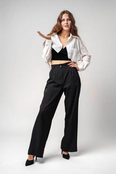 A young woman stands confidently with one hand on her hip and the other extended, dressed in elegant black trousers and a casual chic white shirt, cropped at the waist. Her loose, wavy hair frames her expressive face, and she wears black heeled shoes, completing a modern, fashionable ensemble. The neutral-toned backdrop accentuates her outfit and pose, conveying a sense of poise and style.