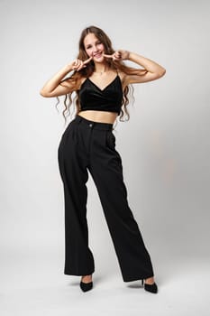 A cheerful young woman with brown hair wearing a black dress is playfully posing against a neutral grey backdrop. She is pointing at her smile, showcasing her happiness, and expressing a positive emotion with sparkling eyes and a beaming expression.