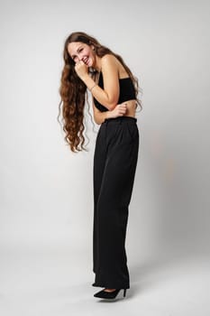 A young woman with long, wavy hair is captured posing expressively with her hand on her cheek and a broad, engaging smile. She is wearing a sleeveless black top which contrasts against a simple, grey backdrop. Her lively expression suggests a moment of genuine happiness or amusement.