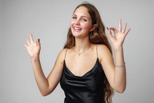 A cheerful young woman with long brown hair is seen giving an okay gesture with both hands, wearing a sleek black dress that suggests a formal or festive occasion. Her joyous expression and the simplicity of the background draw attention to her gesture and attire.