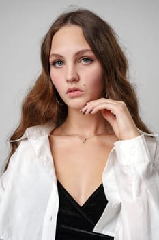 A young woman with long, wavy hair gazes directly at the viewer with a look of confidence. Her delicate touch to her chin, the subtle hues of her natural makeup, and the contrast between her black top and loosely worn white blouse create a portrait of relaxed elegance.