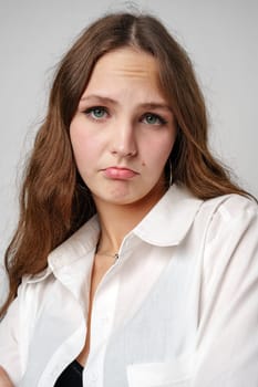 A young woman with long, wavy brown hair and light makeup exhibits a playful yet melancholic pout. She is wearing a casual white button-up shirt, with the top buttons undone, giving the impression of a relaxed or end-of-day mood. The backdrop is plain and grey, drawing full focus to her expressive face.