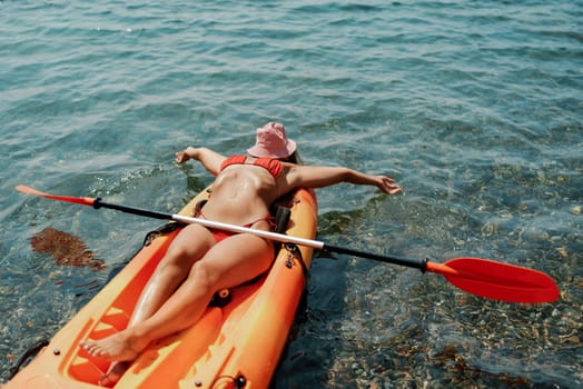 A woman is laying on a kayak in the water. The kayak is orange and has a paddle on it. The woman is wearing a red hat