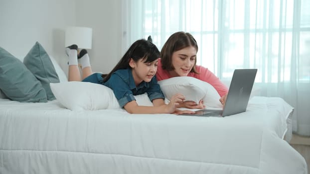 Attractive daughter and caucasian mother watching movie at laptop screen while lie on bad. Happy family spend time together before bed time. Parent and cute girl enjoy playing games together Pedagogy.