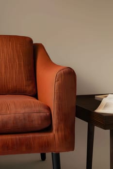 A hardwood orange chair sits comfortably next to a rectangular table with a book on it. The wood stain flooring complements the wood accents